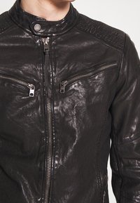Giacca di pelle nera con zip frontale, due tasche sul petto con zip, dettagli a costine sulle spalle e una texture liscia e leggermente lucida.