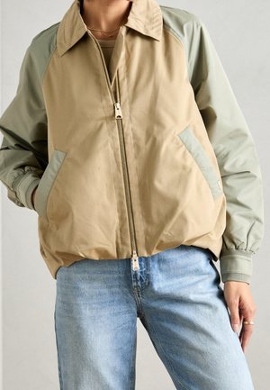 Person iført beige og lysegrøn lynlåstrøje med hænderne i lommerne og lyseblå jeans mod en ensfarvet baggrund.