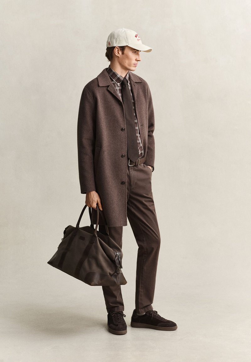 Giovane uomo che indossa un cappotto marrone, una camicia a quadri con cravatta, un berretto bianco, pantaloni e scarpe scure, che tiene una grande borsa da viaggio scura.