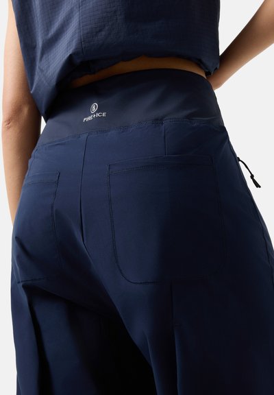 Nahaufnahme einer Person, die marineblaue Hosen mit Gesäßtaschen und einem hohen Taillenbund trägt, auf dem "FIRE+ICE" steht.