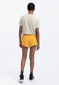 Lichtgrijs t-shirt gecombineerd met gele shorts met een afgeronde zoom. Het model draagt zwarte hoge sneakers met witte accenten.