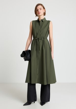 Donna che indossa un vestito midi verde oliva senza maniche con bottoni sopra pantaloni neri a gamba larga, tenendo una pochette nera in pelle piegata.