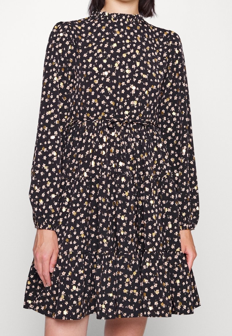 Robe noire à manches longues avec petit imprimé floral rose clair et doré, col montant, taille cintrée et jupe à volants portée par une personne debout.