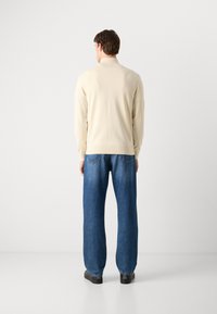 Pull en tricot couleur crème avec un col côtelé, associé à un jeans en denim bleu ample et des chaussures noires, vu de derrière.