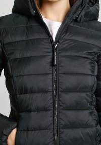 Svart puffjacka med kviltat mönster, huva och framdragkedja. Har en slät, lätt tyg med en glänsande yta.