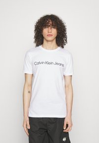 White cotton t-shirt featuring "Calvin Klein Jeans" logo in black text. Short sleeves, crew neck, smooth texture, and casual fit.