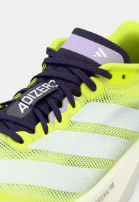 Zapato deportivo amarillo brillante con parte superior de malla, cordones y detalles en púrpura oscuro. Presenta rayas blancas y la marca "ADIZERO" en el lateral.