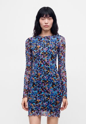 Femme portant une robe ajustée à manches longues avec un motif floral multicolore sur fond blanc uni.