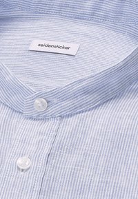 Gros plan sur le col d'une chemise rayée bleu clair et blanc avec des boutons blancs et une étiquette de marque "seidensticker" à l'intérieur du col.