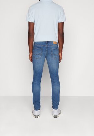 Homme portant un polo bleu clair, un jean bleu ajusté et des baskets blanches, debout face à un mur blanc uni.