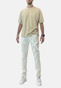 T-shirt in cotone beige a maniche corte, abbinata a pantaloni di denim bianchi. Il modello indossa sneakers bianche e un orologio nero. Design semplice.