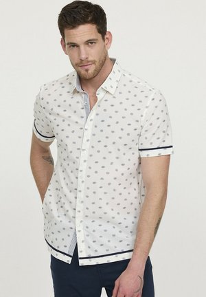 Chemise à manches courtes en blanc avec des pois bleu marine, col intérieur à rayures contrastantes et accents bleu marine aux poignets et à l'ourlet.