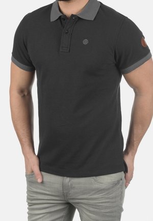 Hombre que lleva una camisa polo negra de manga corta con cuello y puños grises, combinada con vaqueros gris claro, manos en los bolsillos.