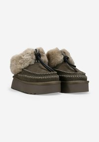 Zapatillas de plataforma de ante marrón con suelas de goma gruesas, forro interior de felpa beige y lazos negros en la parte delantera, vista lateral en ángulo.