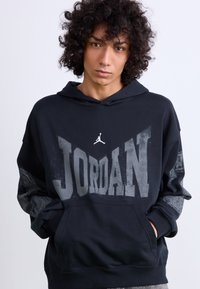 Sort hættejakke med "JORDAN" trykt i store grå bogstaver, der har et lille Jumpman-logo. Stoffet ser blødt ud med en afslappet pasform.