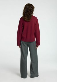 Bordeaux gestrickter Pullover mit abgesenkten Schultern, kombiniert mit grauen, weit geschnittenen Hosen, die ein dezentes vertikales Streifenmuster aufweisen.