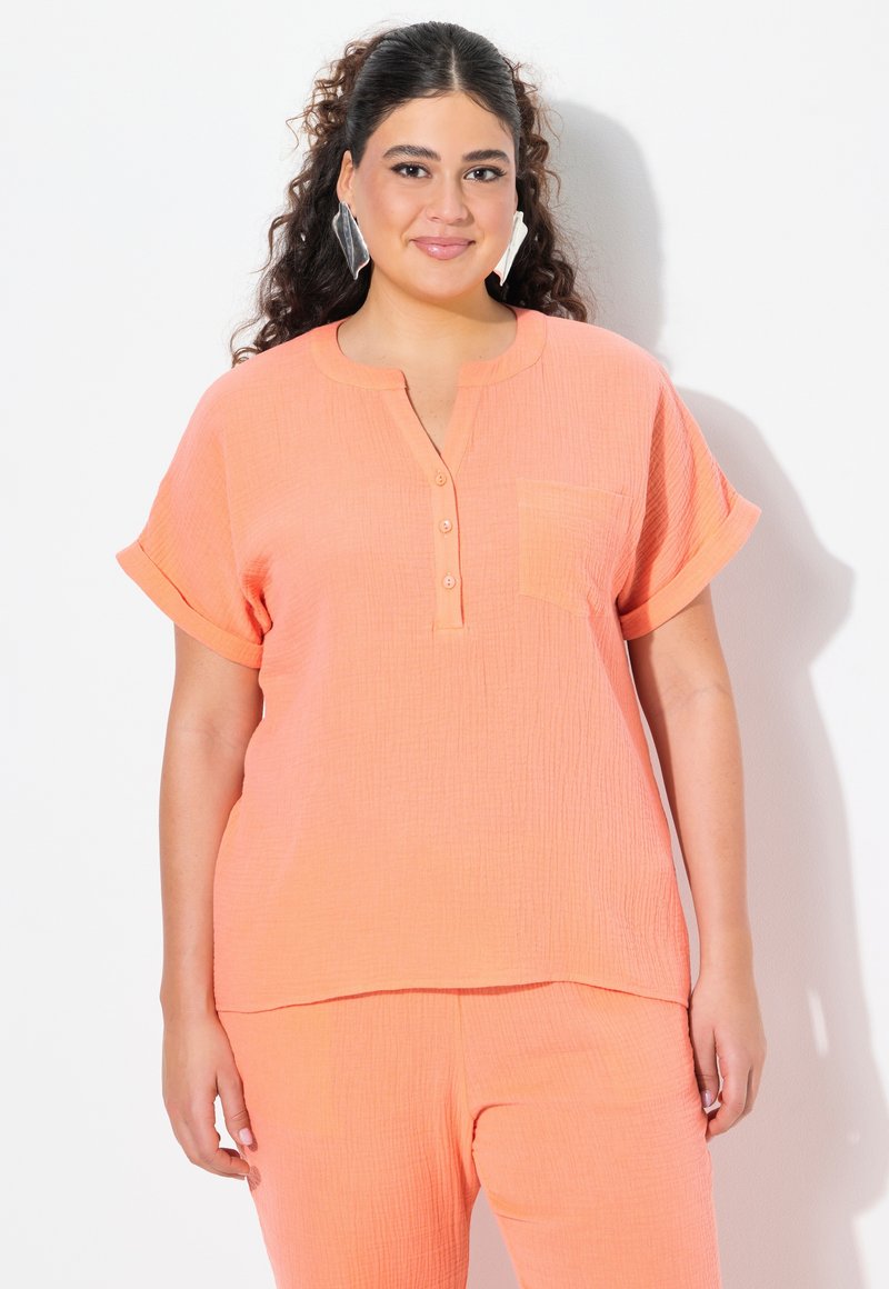 Blusa button-up color pesca, con texture, tasca sul petto e maniche corte arrotolate, abbinata a pantaloni coordinati.