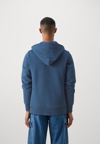 Sudadera de algodón azul con capucha ajustable y puños acanalados, combinada con pantalones de mezclilla de corte relajado. La tela tiene una textura suave.