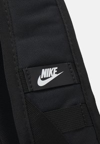 Juodas nailono diržas su tekstūruota paviršiumi, su stačiakampiu Nike logotipo žymeniu baltame fone juodoje aplinkoje, apibrėžtu siuvinėjimu.