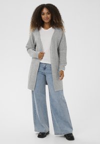 Szürke kötött kardigán nyitott elülső résszel, bordázott ujjaival és laza szabásával, világoskék bőszárú farmerrel és fekete cipővel párosítva.