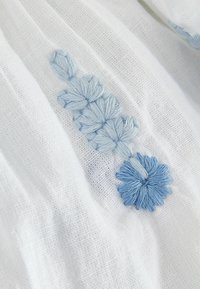 Weißer Stoff mit aufwendiger blauer Stickerei, die florale Muster mit unterschiedlichen Farbtönen zeigt und eine strukturierte, weiche Erscheinung hat.