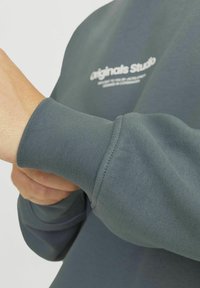 Tamno sivi dukserica s rebrastim manžetama; sadrži otisnuti logo u bijeloj boji. Čini se da je materijal mekan s glatkom teksturom.