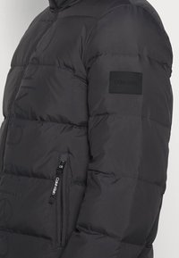 Svart pufferjacka med synlig Calvin Klein-logotyp på ärmplåst och märkt dragkedja på framfickan.