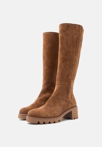 Pár vysokých hnědých semišových bot na blokovém podpatku s drážkovanou podrážkou, které mají vertikální prošívání po zadních stranách.