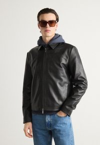 Black leather jacket with a collar and zippered front, layered over a gray hoodie. Worn with light blue jeans and brown sunglasses.