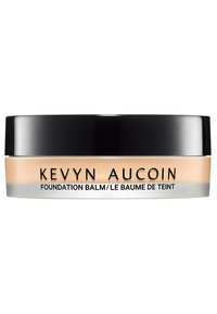Baume de la fondation Kevyn Aucoin dans un pot transparent avec un couvercle noir. Le baume a une teinte beige et une surface lisse. Détails argentés sur la base.