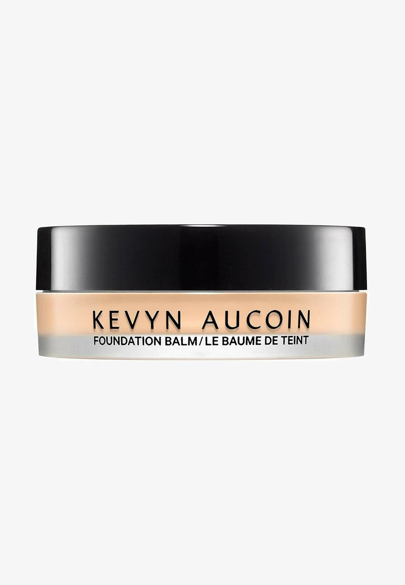 Balsamo della Kevyn Aucoin Foundation in un barattolo trasparente con un coperchio nero. Il balsamo ha una tonalità beige e una superficie liscia. Dettagli in argento sulla base.