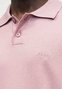 Κοντινό πλάνο ενός ατόμου που φοράει ανοιχτό ροζ πόλο μπλούζα με δύο κουμπιά και το κέντημα «BOSS» στο στήθος.
