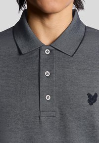 Grå polo t-shirt med tekstureret stof, krave, fire knaplukninger og et mørkeblåt broderet logo på brystet.