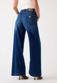 Jeans de mezclilla azul de pierna ancha con una cintura alta, que presentan un parche de cuero en la parte trasera, dos bolsillos traseros y una textura suave.
