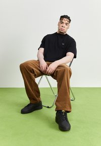 Svart t-shirt med liten logotyp, bruna manchesterbyxor och svarta sneakers. Modellen sitter på en metallstol mot en ljus bakgrund.