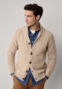 Man in een beige gebreide cardigan over een blauwe button-up shirt en bruine broek, kijkend naar links met handen in de zakken.