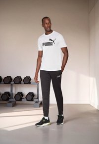 Witte t-shirt met korte mouwen en zwart "PUMA" logo, gecombineerd met zwarte joggingbroek met een klein logo, en zwarte sportschoenen met groene accenten.