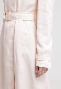 Jasnoróżowy płaszcz trencz z paskiem w talii, długimi rękawami i widoczną boczną kieszenią. Tkanina ma gładką, błyszczącą teksturę.