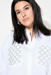 Camicia bianca con bottoni, dotata di due grandi tasche con borchie argentate disposte in un motivo diagonale. Tessuto morbido, vestibilità rilassata e colletto classico.