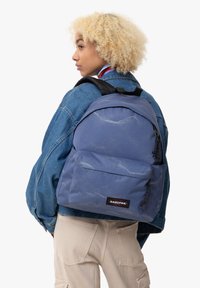 Sac à dos bleu avec tissu texturé, poche avant et bretelles noires. Patch de logo visible sur le bas à l'avant. Porté par une personne en veste en jean.