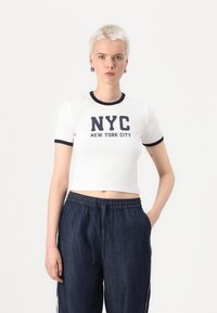 Bijela skraćena majica s mornarskoplavim detaljima oko vrata i ispisom "NYC NEW YORK CITY" u mornarskoplavoj boji. Uparena s tamnoplavim trapericama s vezicama.