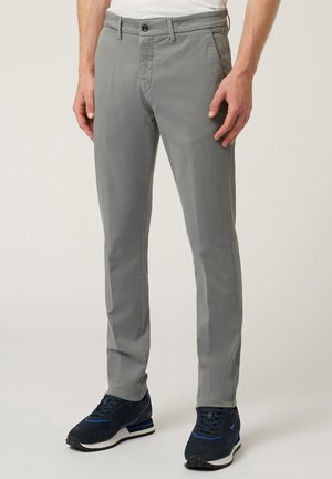 Man draagt lichtgrijze slim-fit broek en donkerblauwe sneakers, staand tegen een effen lichte achtergrond.