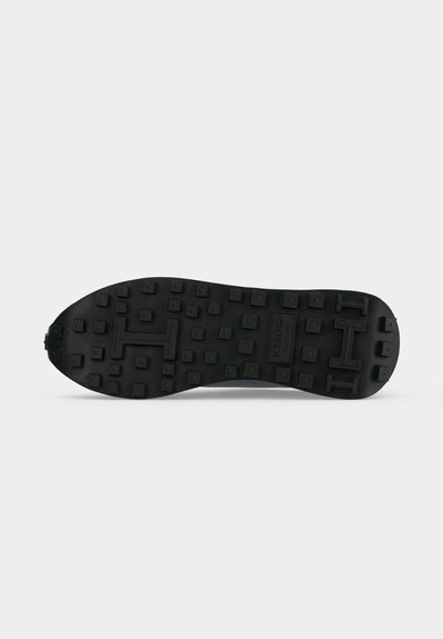 Semelle de chaussure en caoutchouc noir avec des crampons carrés en relief et de grands motifs en forme de lettre H sur un fond blanc uni.