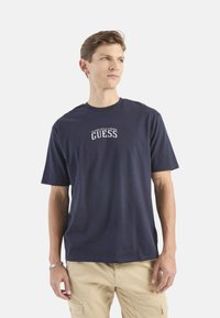 Camiseta de algodón azul marino de manga corta, cuello redondo y logo blanco de "GUESS" impreso en el pecho. Textura suave, corte relajado.
