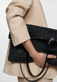 Sac à main noir tissé avec des poignées en cuir lisse. Forme carrée, surface texturée, porté avec une tenue beige clair à fines rayures.