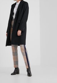 Person klädd i en svart kappa, vit skjorta, grå rutiga byxor med marinblå och orange sidremsor, och svarta klackade ankelstövlar som står inomhus.