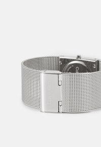 Bracelet de montre en maille argentée avec un boîtier rectangulaire en acier inoxydable. Il est doté d'un fermoir lisse et de l'inscription "Calvin Klein" gravée sur la fermeture.