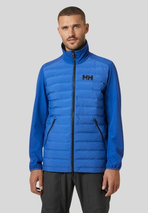 Chaqueta acolchada azul con costuras horizontales, cuello alto, cremallera en la parte frontal y bolsillos laterales. Logo negro en el pecho. Tejido ligero, textura suave.