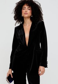 Schwarzer Samtblazer mit tiefem V-Ausschnitt, strukturierten Revers, zwei Fronttaschen und geknöpften Ärmelabschlüssen. Kombiniert mit einer kleinen Clutch.