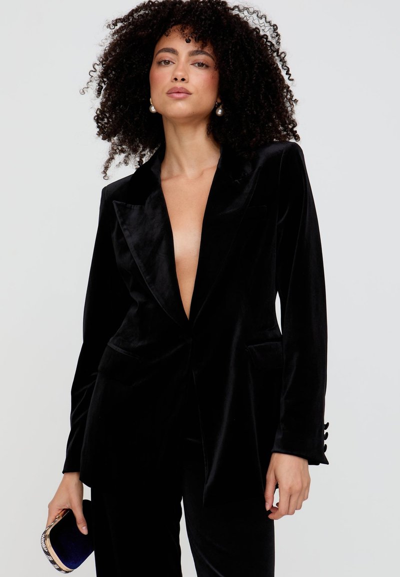Schwarzer Samtblazer mit tiefem V-Ausschnitt, strukturierten Revers, zwei Fronttaschen und geknöpften Ärmelabschlüssen. Kombiniert mit einer kleinen Clutch.